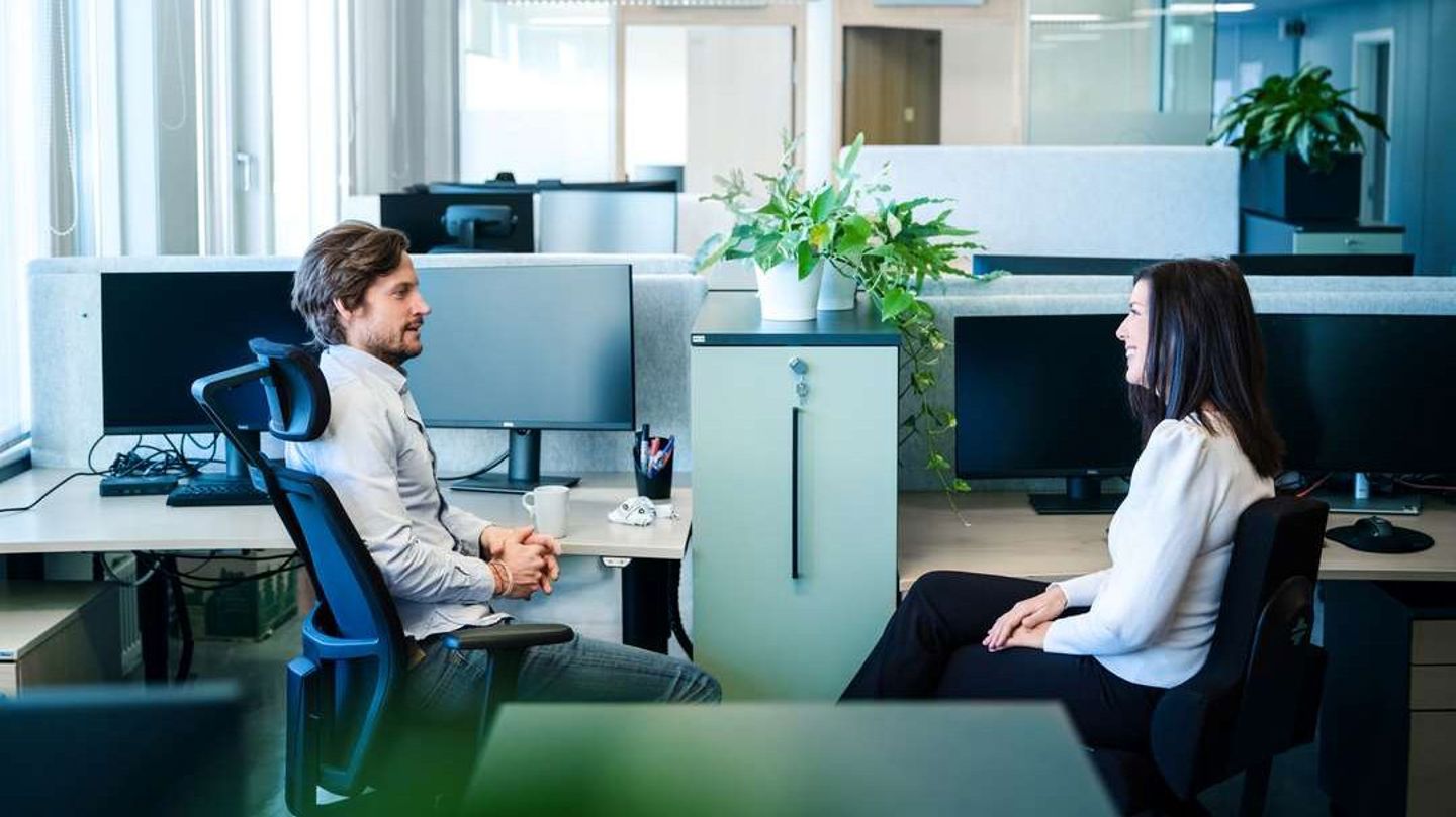 Bildet viser ei kvinne og en mann som snakker med hverandre i et kontorlandskap.