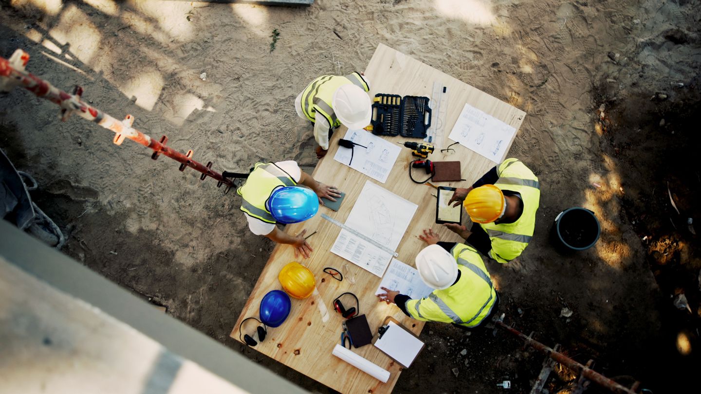 Fire arbeidere med hjelmer på en byggeplass sett ovenfra står rundt et bord med byggetegninger